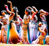 A group of dancers in colorful, flowing costumes perform an intricate dance routine on stage, each striking elegant and expressive poses. The costumes feature detailed embellishments and a harmonious blend of colors, contributing to a sense of movement and grace.