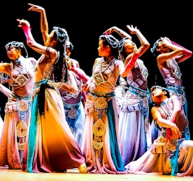 A group of dancers in colorful, flowing costumes perform an intricate dance routine on stage, each striking elegant and expressive poses. The costumes feature detailed embellishments and a harmonious blend of colors, contributing to a sense of movement and grace.
