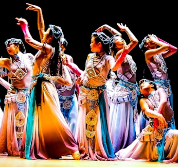 A group of dancers in colorful, flowing costumes perform an intricate dance routine on stage, each striking elegant and expressive poses. The costumes feature detailed embellishments and a harmonious blend of colors, contributing to a sense of movement and grace.