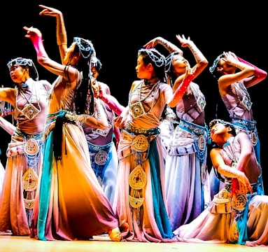 A group of dancers in colorful, flowing costumes perform an intricate dance routine on stage, each striking elegant and expressive poses. The costumes feature detailed embellishments and a harmonious blend of colors, contributing to a sense of movement and grace.