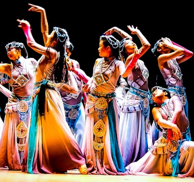 A group of dancers in colorful, flowing costumes perform an intricate dance routine on stage, each striking elegant and expressive poses. The costumes feature detailed embellishments and a harmonious blend of colors, contributing to a sense of movement and grace.