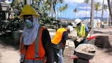 Several construction workers are wearing safety gear, including hard hats, face masks, and reflective vests, standing amidst a construction site with stacks of bricks and palm trees visible. In the background, a scenic view of mountains and a clear blue sky can be seen.