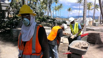 Several construction workers are wearing safety gear, including hard hats, face masks, and reflective vests, standing amidst a construction site with stacks of bricks and palm trees visible. In the background, a scenic view of mountains and a clear blue sky can be seen.