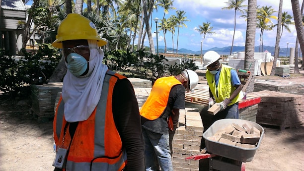 Several construction workers are wearing safety gear, including hard hats, face masks, and reflective vests, standing amidst a construction site with stacks of bricks and palm trees visible. In the background, a scenic view of mountains and a clear blue sky can be seen.