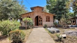 A quaint wooden chapel with arched windows surrounded by lush greenery and bright blue skies. A stone cross stands in the foreground amidst colorful bushes and trees lining a concrete path leading to the chapel entrance.