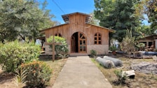 A quaint wooden chapel with arched windows surrounded by lush greenery and bright blue skies. A stone cross stands in the foreground amidst colorful bushes and trees lining a concrete path leading to the chapel entrance.