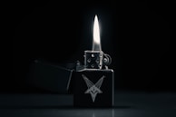 A metallic lighter with an intricate design of a goat head is open with a prominent flame. The background is dark, emphasizing the lighter and the flame.