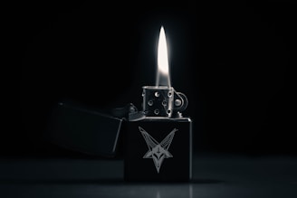 A metallic lighter with an intricate design of a goat head is open with a prominent flame. The background is dark, emphasizing the lighter and the flame.