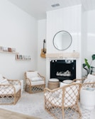 A cozy living room with a modern, minimalist design featuring wicker chairs with white cushions arranged around a fireplace. Above the fireplace is a round mirror and a string of small lights. A guitar is mounted on the wall beside a couple of shelves displaying small books or artwork. A large green plant adds a touch of nature to the room. The ceiling is paneled and the walls are painted white, contributing to a bright and airy atmosphere.
