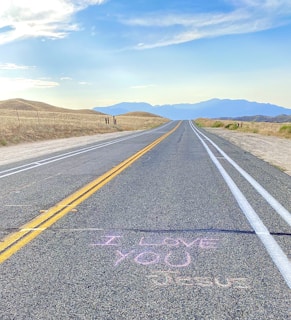 A deserted road stretches into the horizon, with a message written in chalk on the asphalt reading 'I love you Jesus.' The surrounding landscape is dry and grassy, with distant mountains visible under a clear blue sky.