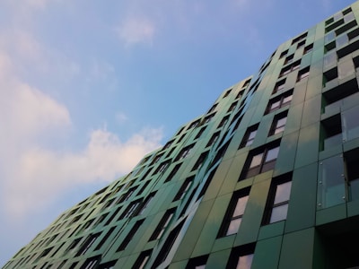 A modern building with a facade of green panels and large windows reaches towards the sky at an angle. The building’s surface reflects light, and the clear blue sky with scattered clouds serves as a backdrop.