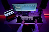 An individual is sitting at a desk in a dimly lit room with a vibrant red and blue ambient lighting. The person is working on a computer setup that includes a large monitor displaying a complex application with various tracks or data lines. There is also a laptop on the desk displaying a webpage. The desk is filled with tech accessories, including speakers, a mouse, a keyboard with backlighting, and some cables.