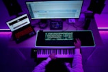 A person sits at a desk with a music production setup. There are two monitors, one displaying a digital audio workstation and another with a browser open. A keyboard and mouse sit on a glowing desk mat, and a MIDI keyboard is in front of the person. The ambient lighting casts a purple hue over the scene.
