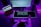 A person sits at a desk with a music production setup. There are two monitors, one displaying a digital audio workstation and another with a browser open. A keyboard and mouse sit on a glowing desk mat, and a MIDI keyboard is in front of the person. The ambient lighting casts a purple hue over the scene.