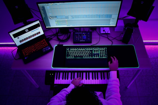 A person sits at a desk with a music production setup. There are two monitors, one displaying a digital audio workstation and another with a browser open. A keyboard and mouse sit on a glowing desk mat, and a MIDI keyboard is in front of the person. The ambient lighting casts a purple hue over the scene.