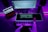A person sits at a desk with a music production setup. There are two monitors, one displaying a digital audio workstation and another with a browser open. A keyboard and mouse sit on a glowing desk mat, and a MIDI keyboard is in front of the person. The ambient lighting casts a purple hue over the scene.