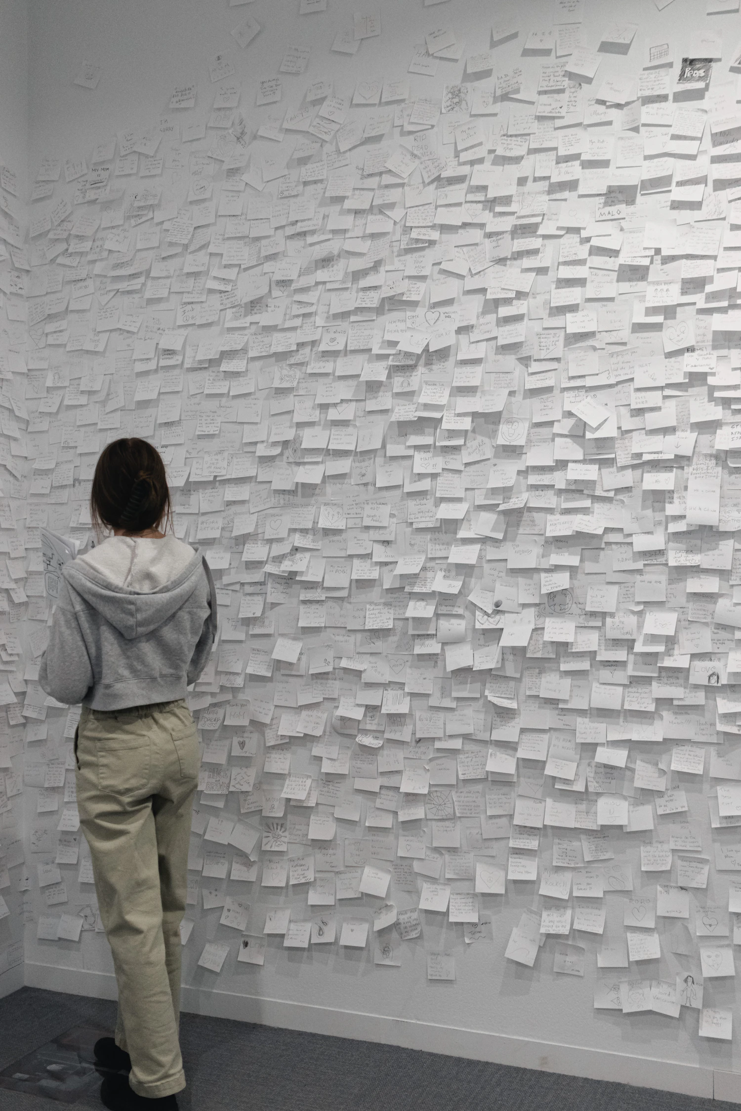 A woman standing at the foot of a white wall covered from floor to ceiling with thousands of small pinned paper notes, dwarfed by it