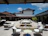An outdoor courtyard with wicker seating and white cushions surrounds a central stone fountain. The area is framed by buildings with red tile roofs against a clear blue sky. Potted plants add a touch of greenery.