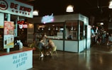A cozy indoor food court setting featuring various food stands including a coffee stand named 'Be Kind Coffee' and a salad stand called 'The Salad Stand'. There are people sitting at a table eating and chatting. The environment is warmly lit with hanging lights and has a casual, inviting atmosphere. Other food stalls and a gathering of people can be seen in the background.