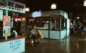 A cozy indoor food court setting featuring various food stands including a coffee stand named 'Be Kind Coffee' and a salad stand called 'The Salad Stand'. There are people sitting at a table eating and chatting. The environment is warmly lit with hanging lights and has a casual, inviting atmosphere. Other food stalls and a gathering of people can be seen in the background.