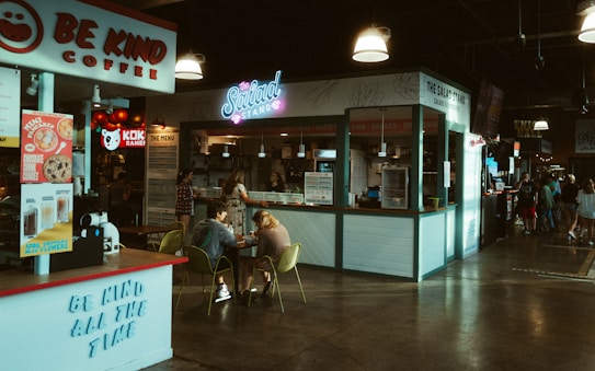 A cozy indoor food court setting featuring various food stands including a coffee stand named 'Be Kind Coffee' and a salad stand called 'The Salad Stand'. There are people sitting at a table eating and chatting. The environment is warmly lit with hanging lights and has a casual, inviting atmosphere. Other food stalls and a gathering of people can be seen in the background.