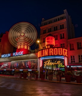 Bright red neon lights illuminate the iconic windmill facade of a famous cabaret, capturing the bustling night scene. The architectural details appear vibrant against the deep blue night sky, enhancing the allure of the venue. A few pedestrians can be seen on the quiet street, lined with potted plants and barriers.