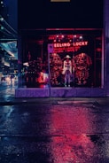 A nighttime city scene features a storefront with a mannequin wearing a hat and clothing, illuminated by red neon lights that reflect on the wet pavement. The words 'FEELING LUCKY' glow in neon above the display.