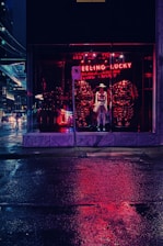 A nighttime city scene features a storefront with a mannequin wearing a hat and clothing, illuminated by red neon lights that reflect on the wet pavement. The words 'FEELING LUCKY' glow in neon above the display.