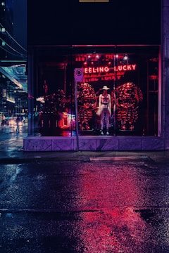 A nighttime city scene features a storefront with a mannequin wearing a hat and clothing, illuminated by red neon lights that reflect on the wet pavement. The words 'FEELING LUCKY' glow in neon above the display.