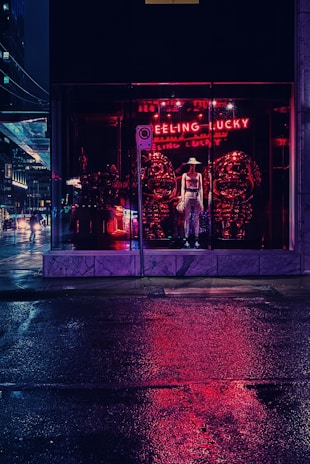 A nighttime city scene features a storefront with a mannequin wearing a hat and clothing, illuminated by red neon lights that reflect on the wet pavement. The words 'FEELING LUCKY' glow in neon above the display.