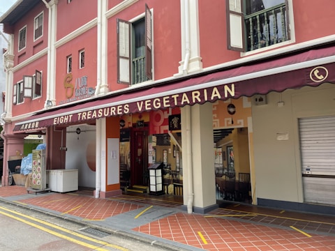 A storefront features a vegetarian restaurant with a maroon and yellow sign that reads 'Eight Treasures Vegetarian'. The building has a traditional architectural style with open windows and columns painted in a brick red color. The entrance is inviting with a menu display and chairs visible inside.
