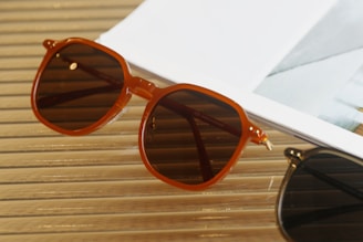 A pair of brown sunglasses with a vintage design rests on a wooden surface next to the corner of a white magazine. The setting exudes a relaxed and stylish vibe.