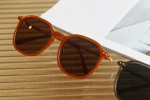 A pair of brown sunglasses with a vintage design rests on a wooden surface next to the corner of a white magazine. The setting exudes a relaxed and stylish vibe.
