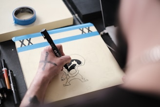 A person is drawing a cartoon-like figure on a notepad using a black marker. The scene includes a roll of blue masking tape and several other markers on a desk. The artist has tattoos on their arm.