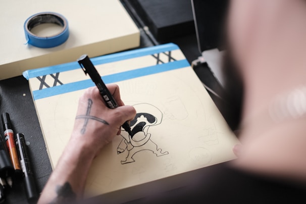 A person is drawing a cartoon-like figure on a notepad using a black marker. The scene includes a roll of blue masking tape and several other markers on a desk. The artist has tattoos on their arm.