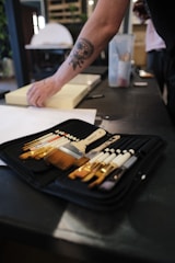 A set of painting brushes is neatly arranged in a black case on a table. A person's arm with a distinctive tattoo is reaching towards a light-colored object on the table. The background includes blurred elements like a large container and some shelving.