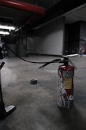 A fire extinguisher with a worn, red surface sits on an industrial-style concrete floor. It has a long black hose connected to it, and there's duct tape on the ground nearby. The setting appears dimly lit, with industrial elements like metal pipes and structural features visible in the background.