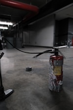 A fire extinguisher with a worn, red surface sits on an industrial-style concrete floor. It has a long black hose connected to it, and there's duct tape on the ground nearby. The setting appears dimly lit, with industrial elements like metal pipes and structural features visible in the background.