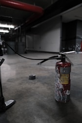 A fire extinguisher with a worn, red surface sits on an industrial-style concrete floor. It has a long black hose connected to it, and there's duct tape on the ground nearby. The setting appears dimly lit, with industrial elements like metal pipes and structural features visible in the background.