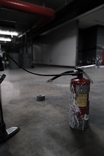 A fire extinguisher with a worn, red surface sits on an industrial-style concrete floor. It has a long black hose connected to it, and there's duct tape on the ground nearby. The setting appears dimly lit, with industrial elements like metal pipes and structural features visible in the background.