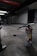A fire extinguisher with a worn, red surface sits on an industrial-style concrete floor. It has a long black hose connected to it, and there's duct tape on the ground nearby. The setting appears dimly lit, with industrial elements like metal pipes and structural features visible in the background.