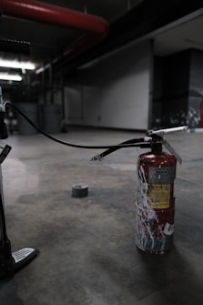 A fire extinguisher with a worn, red surface sits on an industrial-style concrete floor. It has a long black hose connected to it, and there's duct tape on the ground nearby. The setting appears dimly lit, with industrial elements like metal pipes and structural features visible in the background.