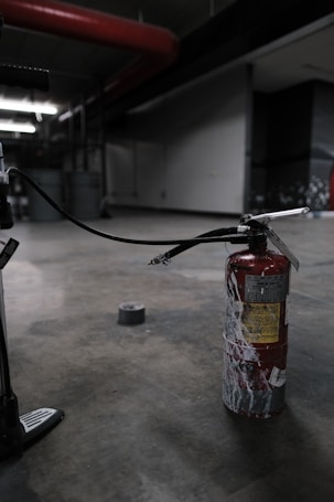 A fire extinguisher with a worn, red surface sits on an industrial-style concrete floor. It has a long black hose connected to it, and there's duct tape on the ground nearby. The setting appears dimly lit, with industrial elements like metal pipes and structural features visible in the background.
