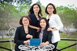 Four women dressed in professional attire are gathered outdoors at a round table. Three of them are standing behind while one is sitting in front holding a tablet. They are all smiling and appear to be enjoying their time together. The background consists of greenery and trees, and the scene seems to be set in a garden or park.