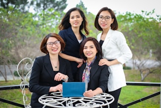 Four women dressed in professional attire are gathered outdoors at a round table. Three of them are standing behind while one is sitting in front holding a tablet. They are all smiling and appear to be enjoying their time together. The background consists of greenery and trees, and the scene seems to be set in a garden or park.