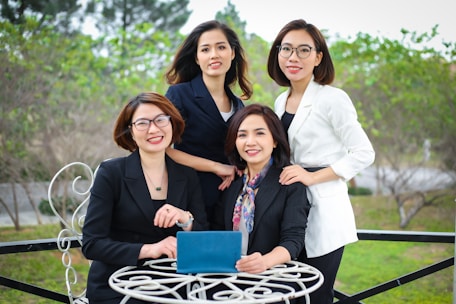 Four women dressed in professional attire are gathered outdoors at a round table. Three of them are standing behind while one is sitting in front holding a tablet. They are all smiling and appear to be enjoying their time together. The background consists of greenery and trees, and the scene seems to be set in a garden or park.
