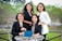 Four women dressed in professional attire are gathered outdoors at a round table. Three of them are standing behind while one is sitting in front holding a tablet. They are all smiling and appear to be enjoying their time together. The background consists of greenery and trees, and the scene seems to be set in a garden or park.