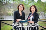 Two women in professional attire are seated outdoors at a white metal table, holding a book and smiling. The background is a blurred view of greenery and trees.