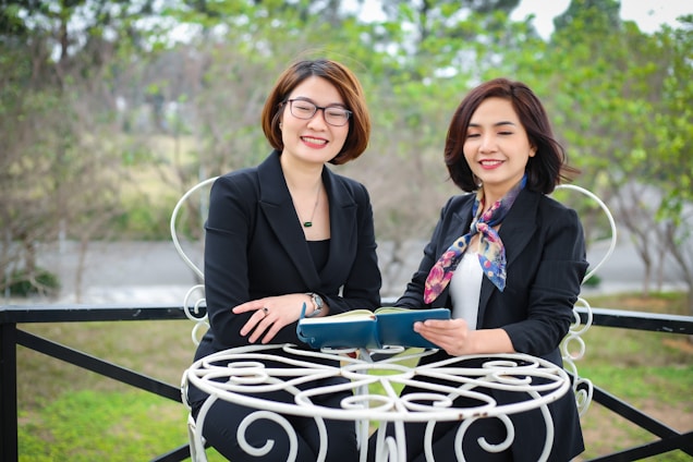Two women in professional attire are seated outdoors at a white metal table, holding a book and smiling. The background is a blurred view of greenery and trees.