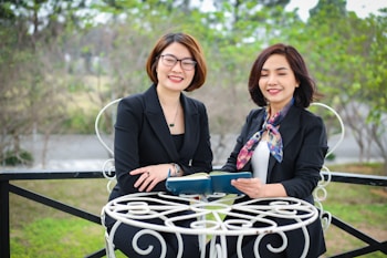 Two women in professional attire are seated outdoors at a white metal table, holding a book and smiling. The background is a blurred view of greenery and trees.